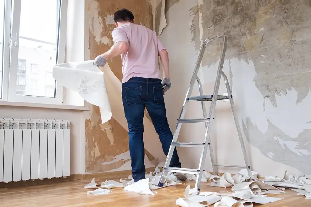 Man verwijdert oud behang van de muur met de hand, naast een trap in een kamer tijdens renovatie.