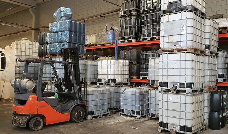 Bij deze chemische groothandel tilt een vorkheftruck gestapelde Intermediate Bulk Containers (IBC's) in een industrieel magazijn. Dit is een veelvoorkomend beeld in de chemische industrie.