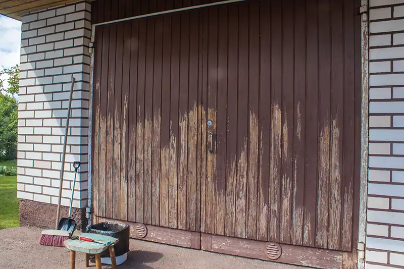 Garagedeuren van hout met gedeeltelijk afgebladderde verf en voorbereide schoonmaakbenodigdheden ernaast.