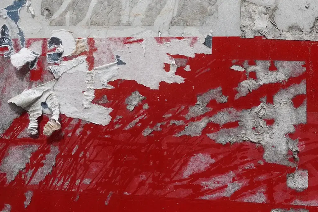 Een close-up afbeelding van hardnekkige lijmresten op een rood oppervlak, met gedeeltelijk afgescheurde en verbleekte lagen van papier en lijm.