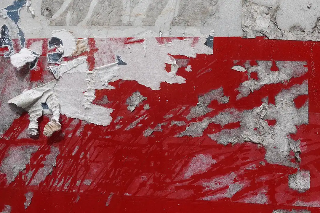 Een close-up afbeelding van hardnekkige lijmresten op een rood oppervlak, met gedeeltelijk afgescheurde en verbleekte lagen van papier en lijm.