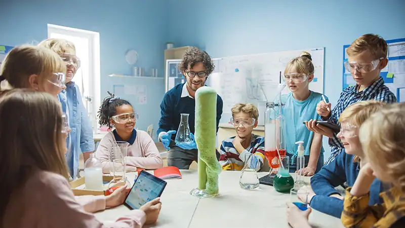 Kinderen dragen veiligheidsbrillen en kijken naar een demonstratie van het olifantentandpasta-experiment in een klaslokaal.